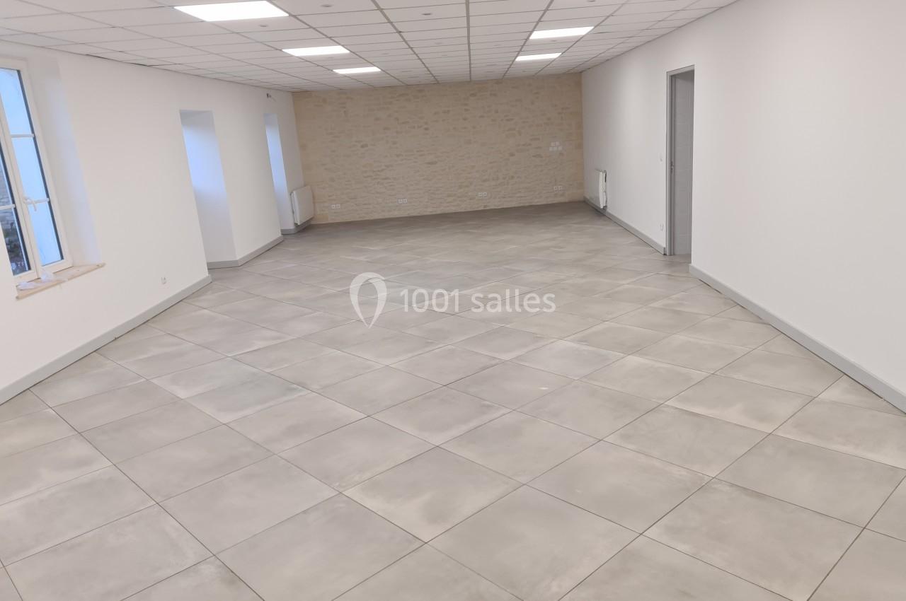 Salle vide avec carrelage gris clair, murs blancs, plafond à dalles et une fenêtre laissant entrer la lumière naturelle.
