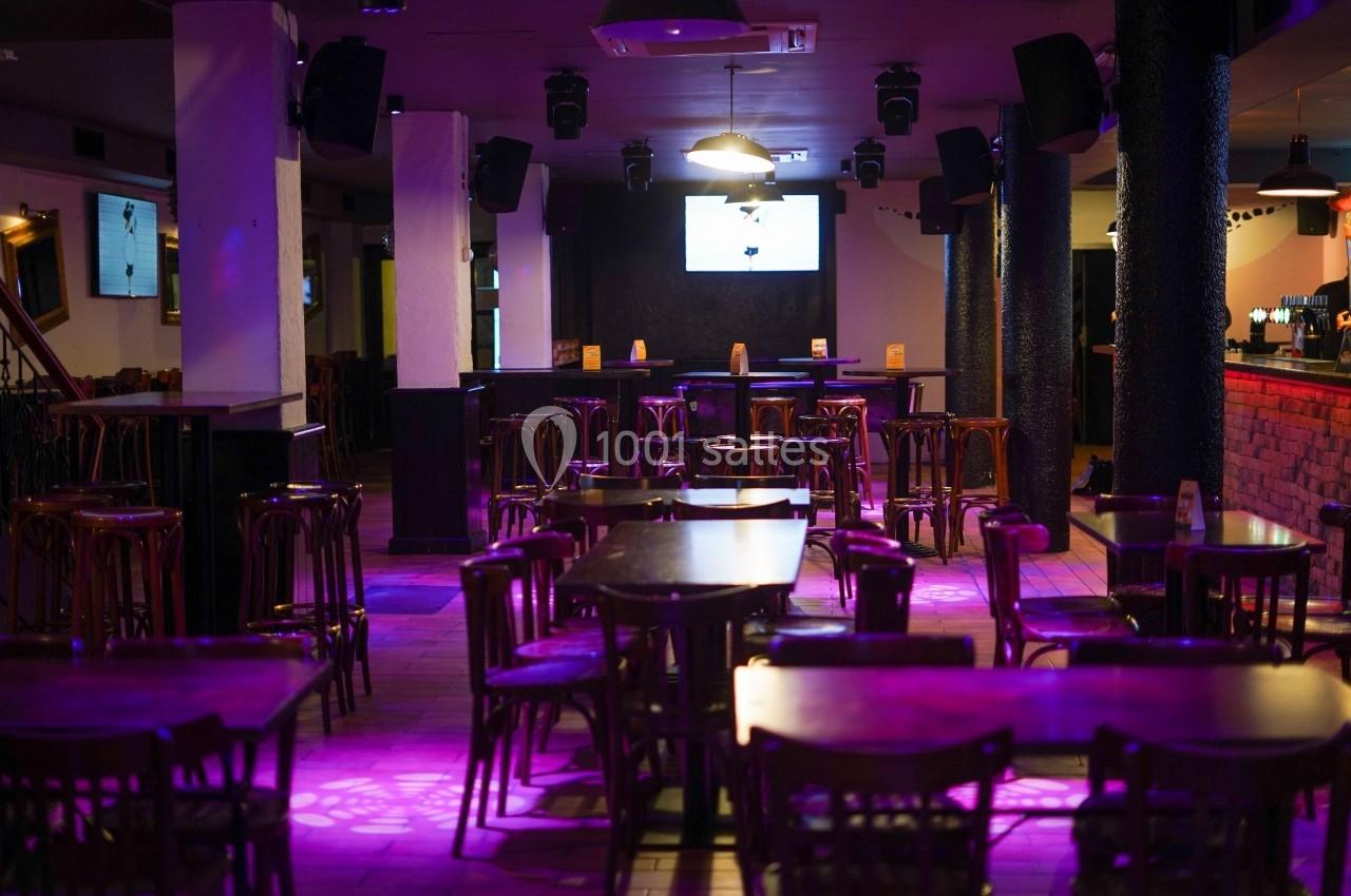 Salle de bar avec tables et chaises en bois, éclairage tamisé violet, écran allumé au fond.