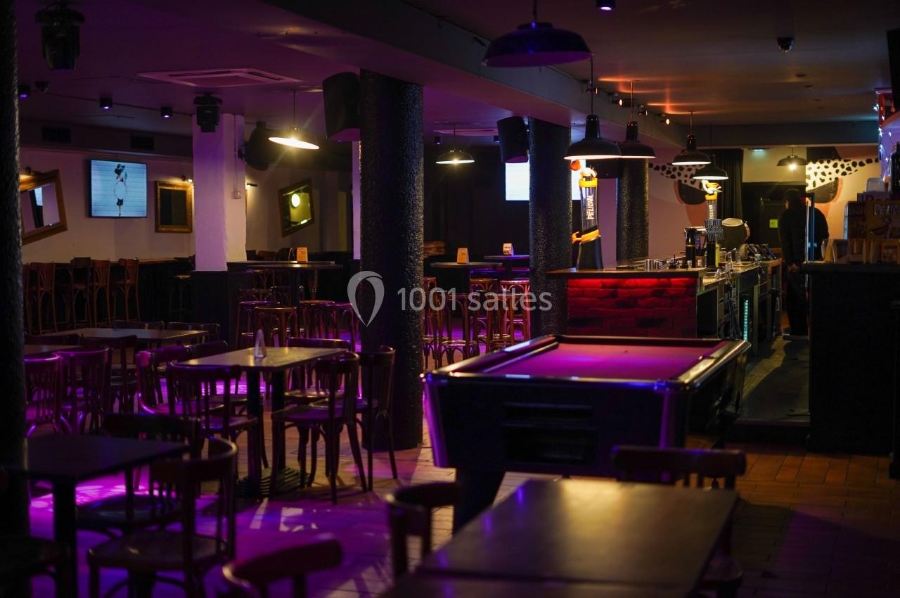 Salle de bar avec tables en bois, billard au premier plan et éclairage tamisé aux tons violets.