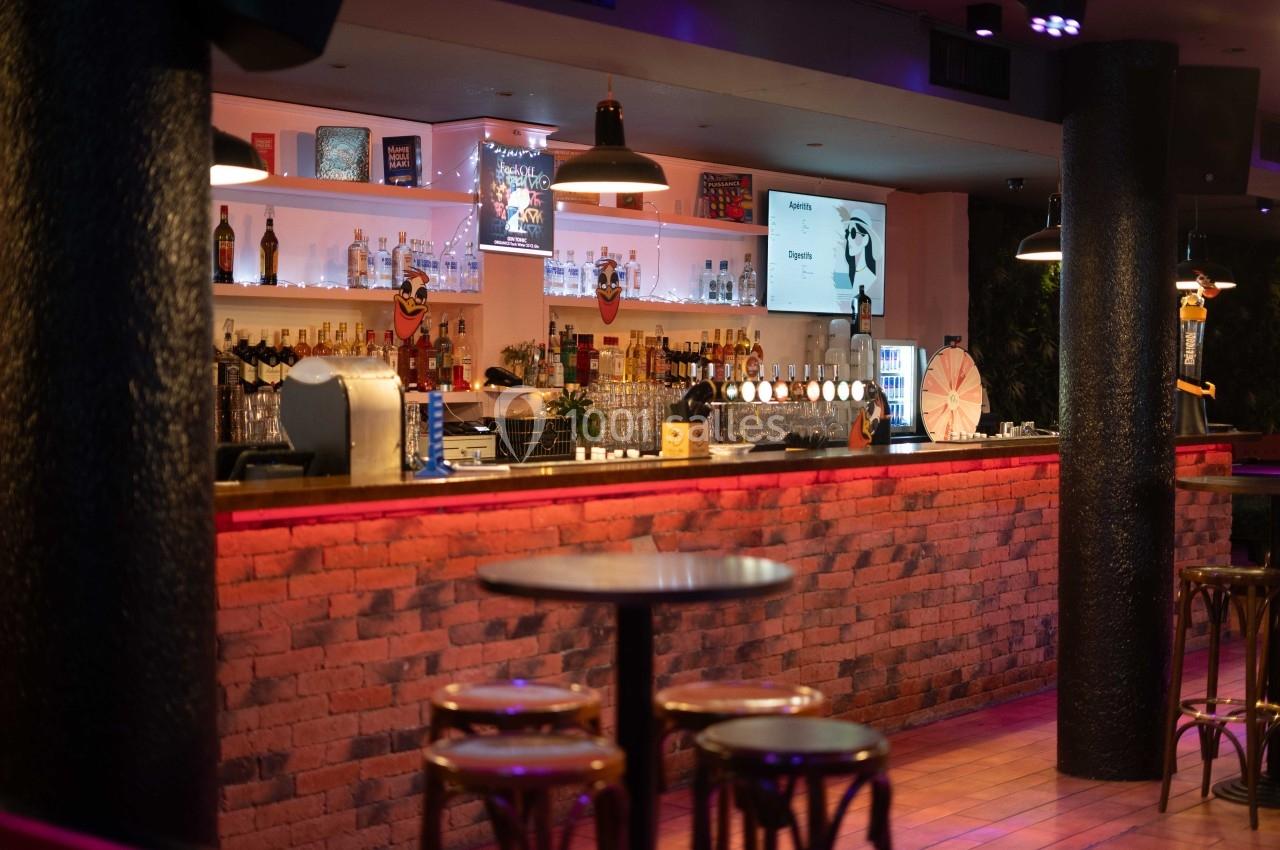 Bar avec comptoir en briques rouges, étagères éclairées contenant des bouteilles, et tables hautes au premier plan.