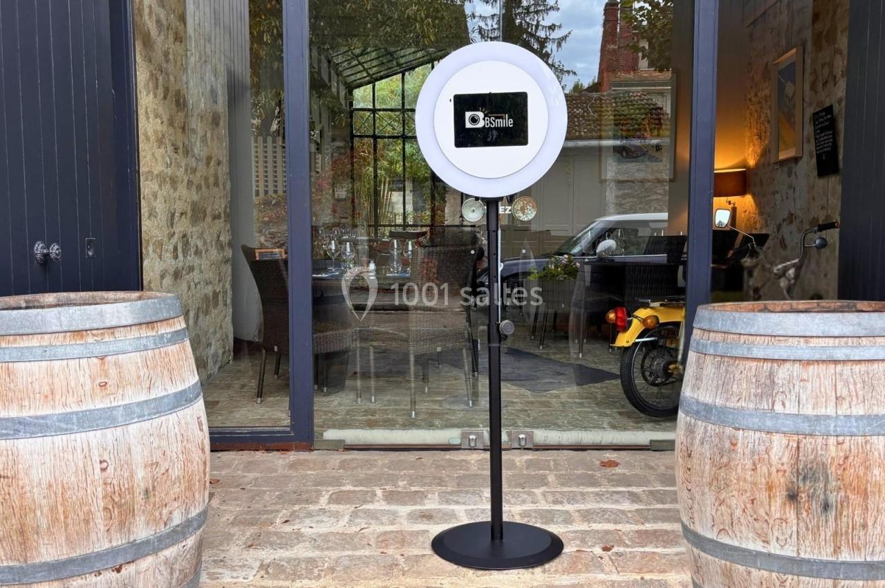 Borne photo avec anneau lumineux placée devant une entrée vitrée, entourée de tonneaux en bois.