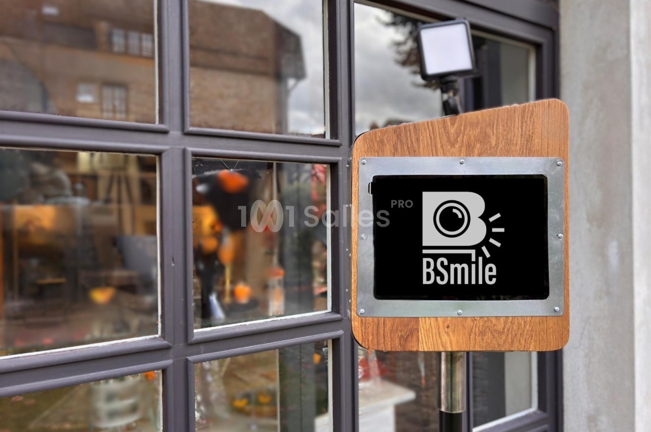 Borne photo en bois avec écran tactile et logo, placée devant une vitrine reflétant des bâtiments et le ciel.