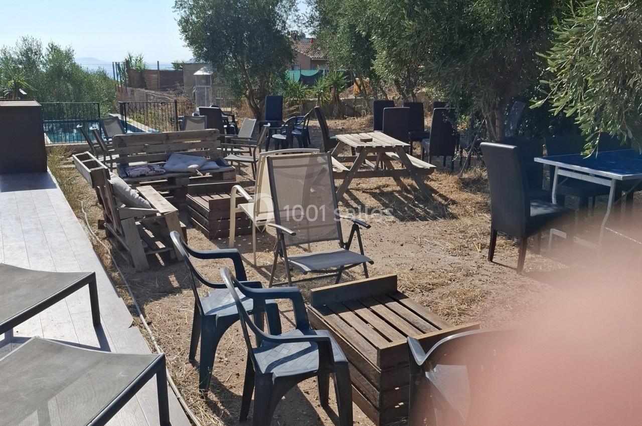 Mobilier de jardin en bois et en plastique disposé sur un sol en terre, entouré d'arbres et proche d'une piscine.