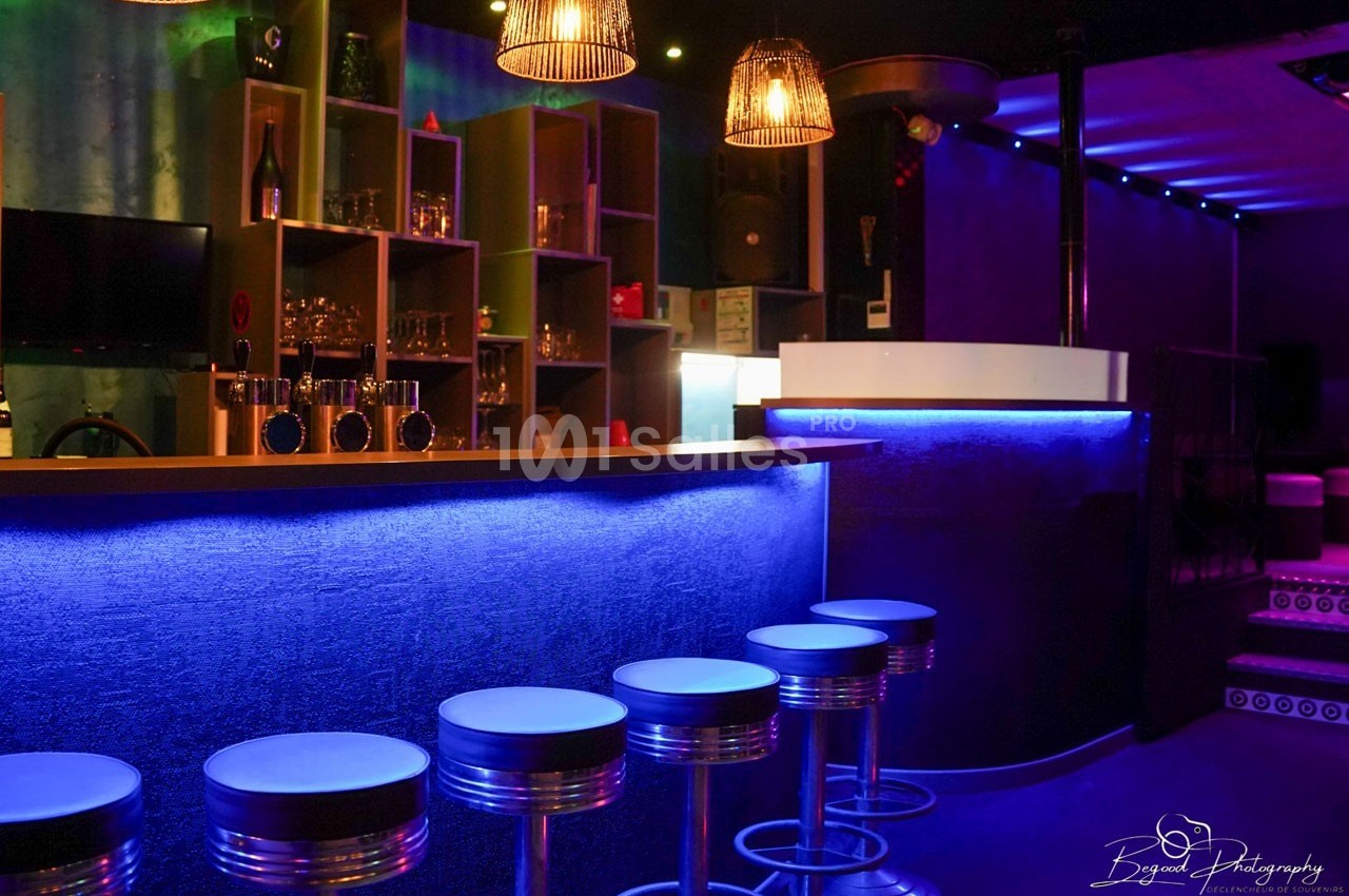 Bar avec comptoir éclairé en bleu, tabourets métalliques alignés et étagères contenant des verres en arrière-plan.