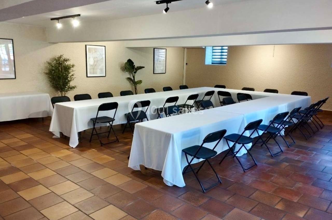 Salle de réunion lumineuse avec tables en U, nappes blanches, chaises noires et sol en carrelage brun.