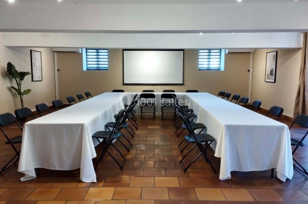 Salle de réunion avec tables en U couvertes de nappes blanches, chaises noires et écran de projection en fond.