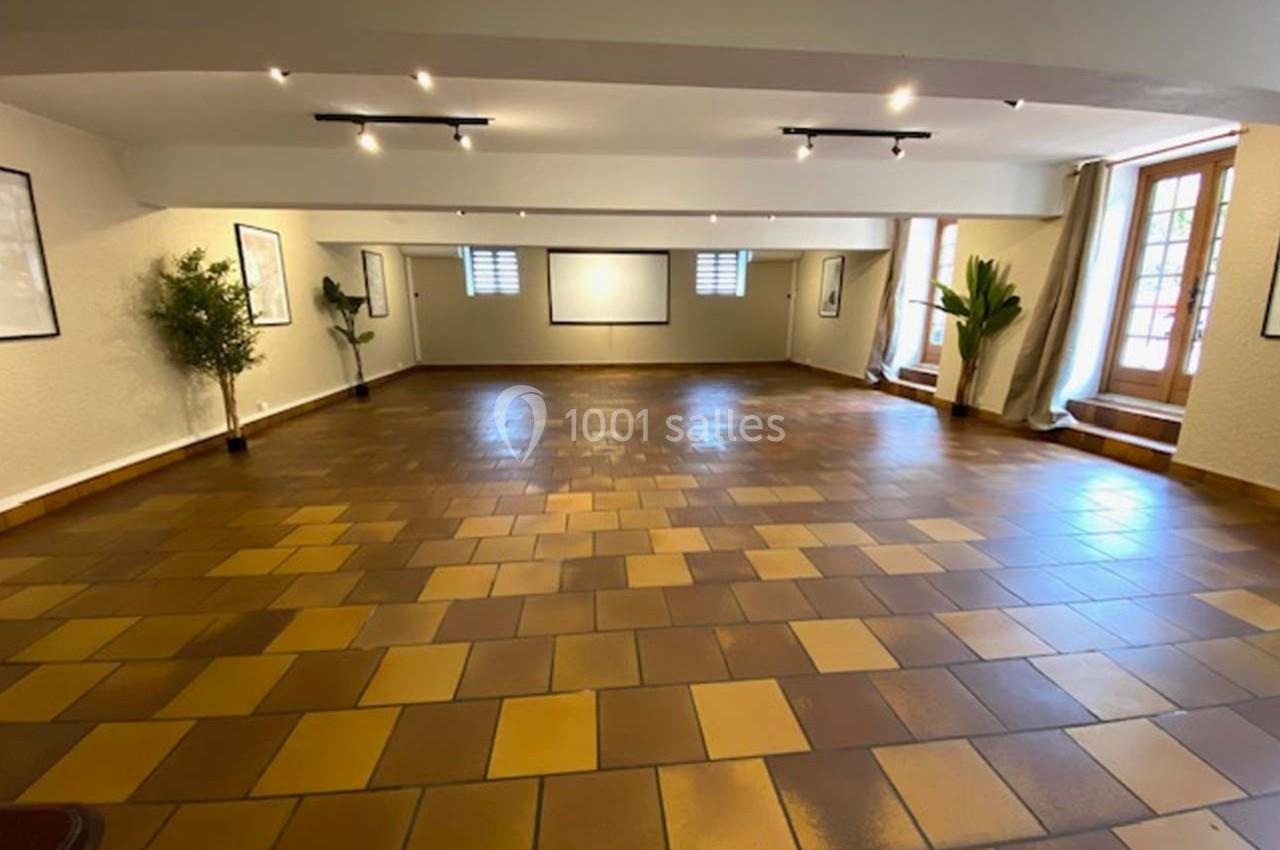 Salle vide avec sol en carrelage marron, murs clairs, grandes fenêtres, plantes et éclairage au plafond.