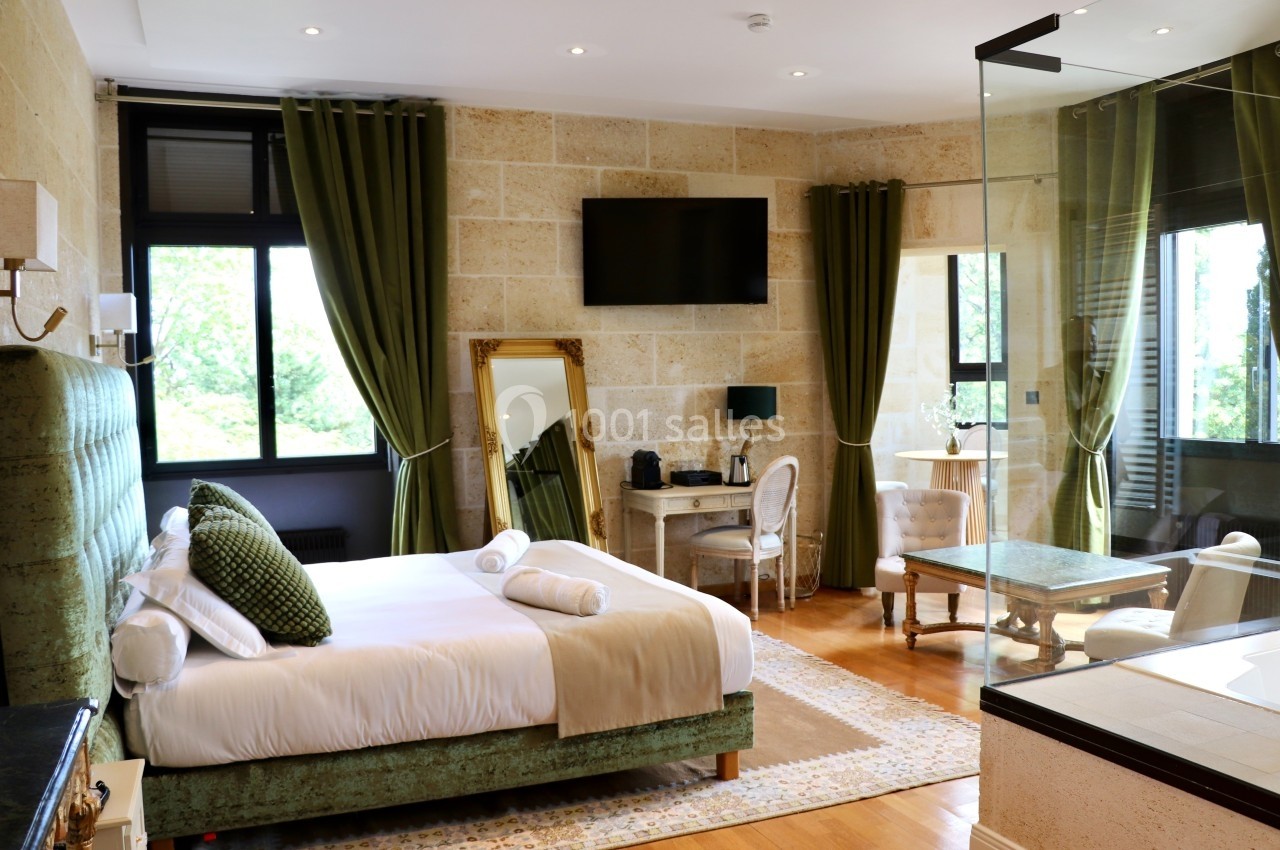Chambre lumineuse avec lit double, fauteuils, bureau, miroir, grandes fenêtres et rideaux verts.