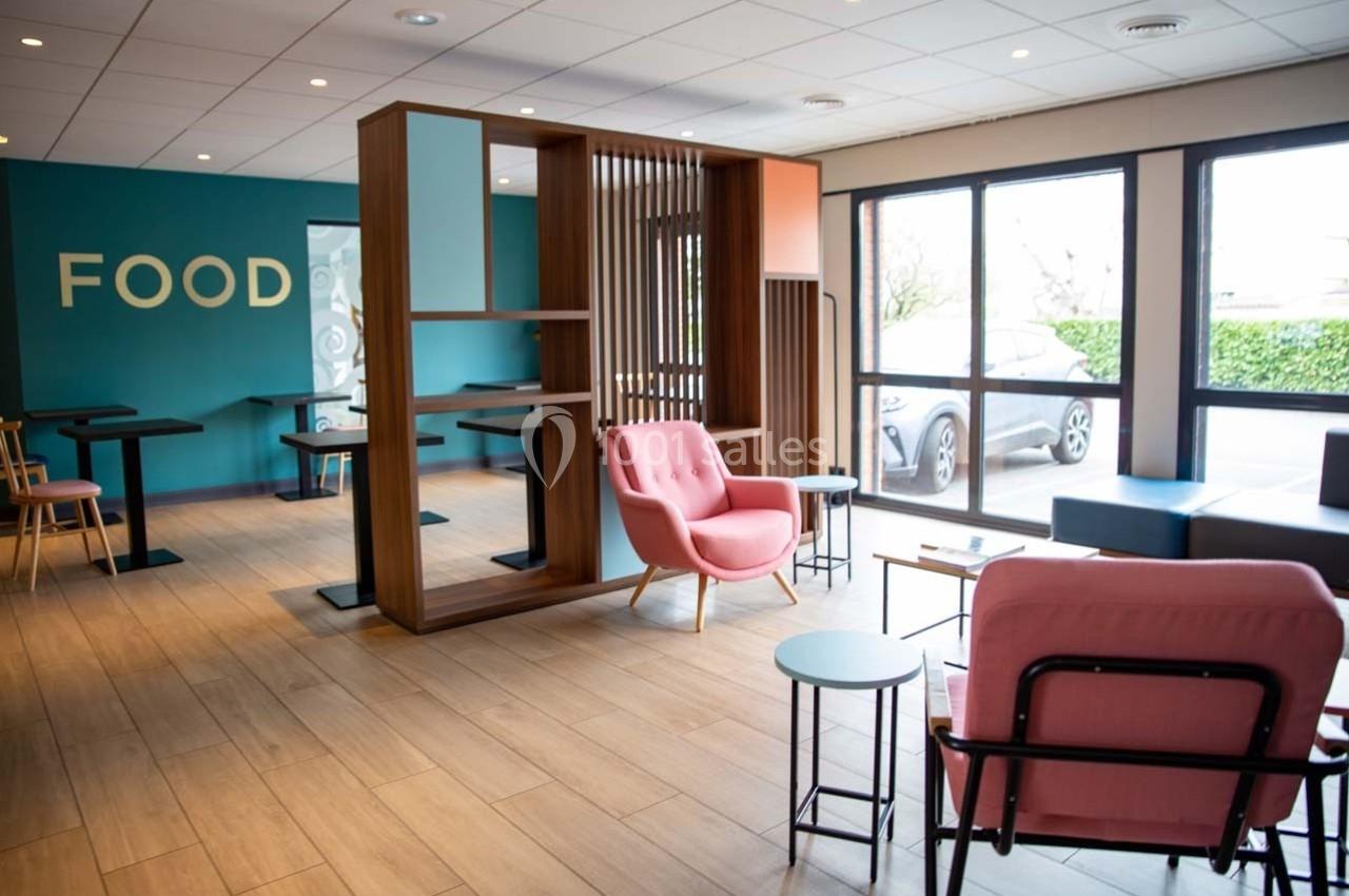 Espace intérieur lumineux avec fauteuils roses, tables basses, cloison en bois et grandes baies vitrées donnant sur l…
