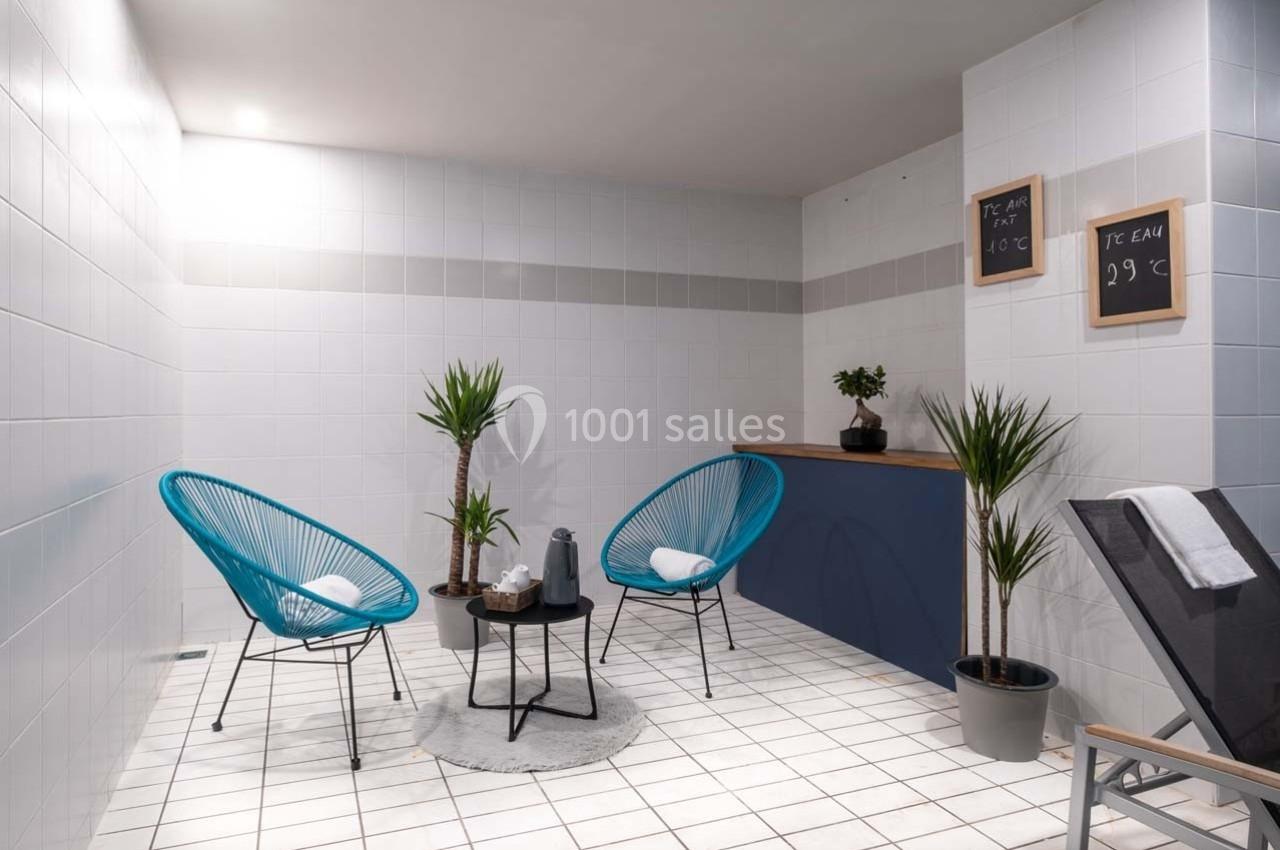 Espace détente avec fauteuils bleus, table basse, plantes et murs carrelés blancs dans une ambiance lumineuse.