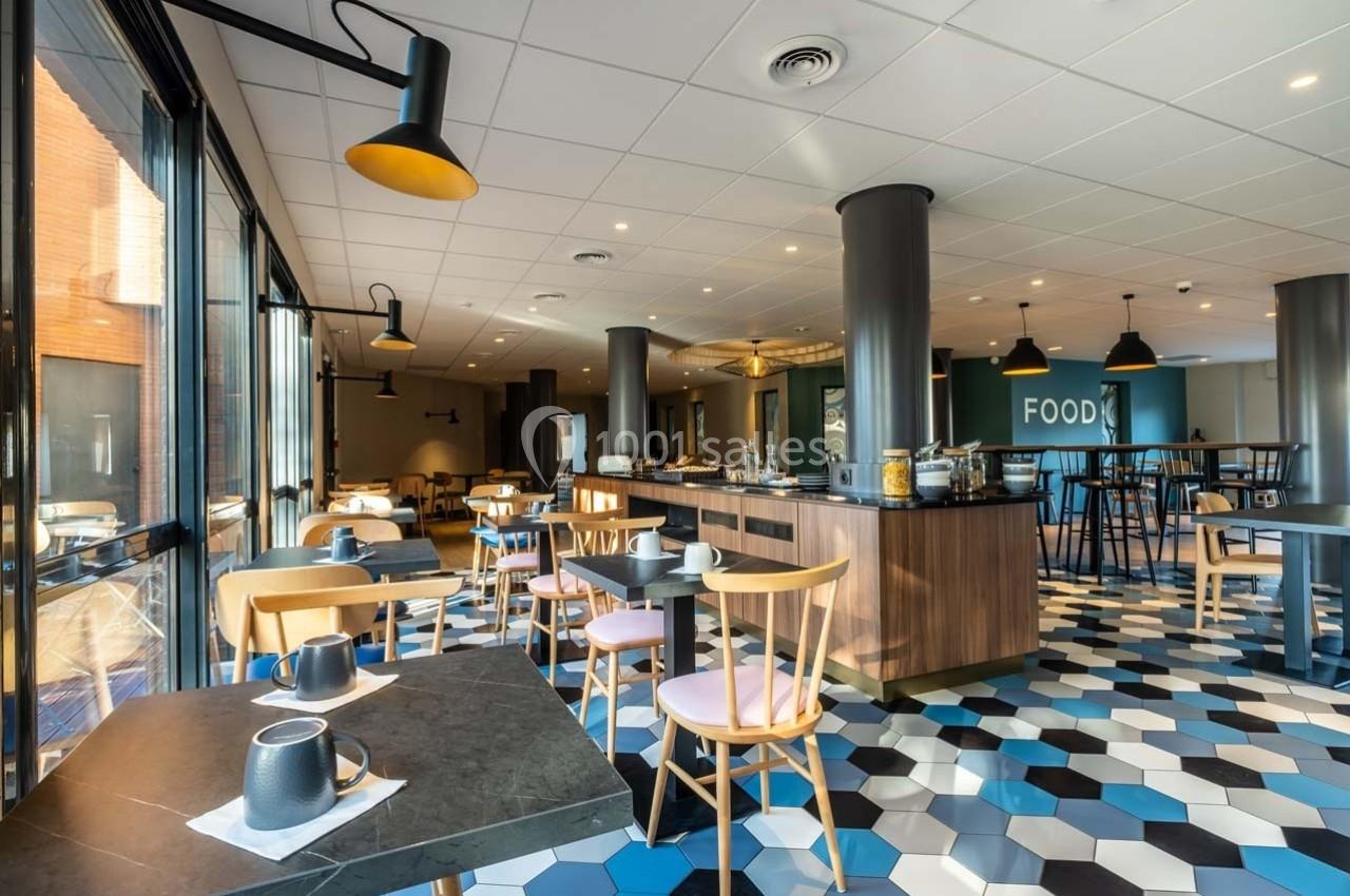 Salle de restaurant lumineuse avec tables, chaises en bois, sol à motifs géométriques et buffet central.