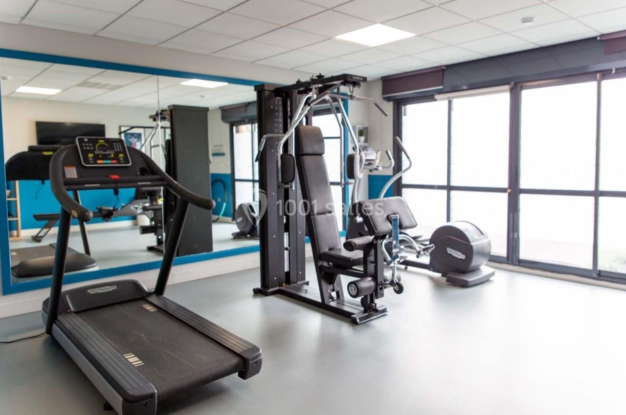 Salle de sport lumineuse avec tapis de course, machine de musculation et grandes baies vitrées.