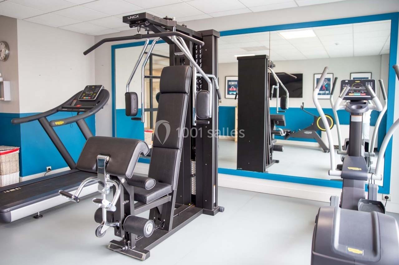 Salle de sport équipée de machines de musculation, tapis de course et vélo elliptique, avec miroir mural.