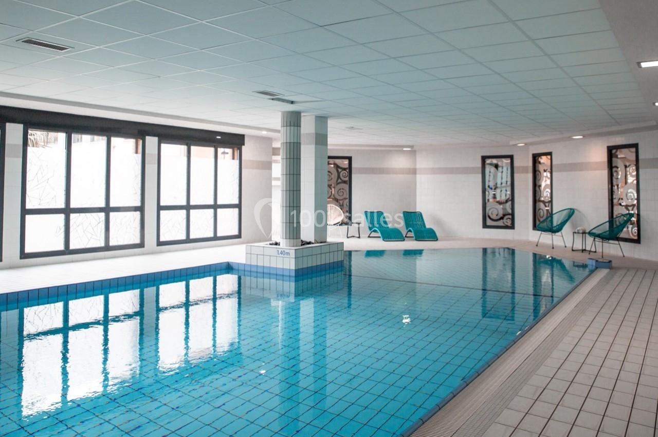 Piscine intérieure avec carrelage bleu, chaises longues et grandes fenêtres laissant entrer la lumière naturelle.