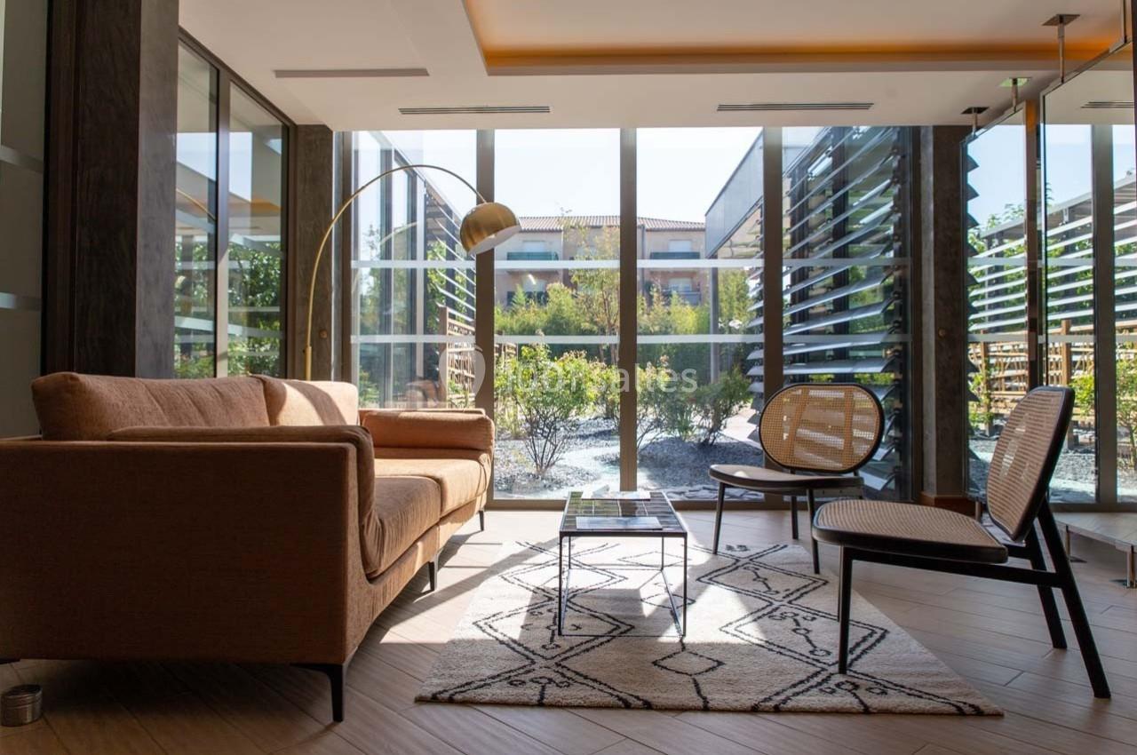 Salon lumineux avec canapé beige, fauteuil en rotin, lampe arquée dorée et grandes baies vitrées donnant sur un jardin.
