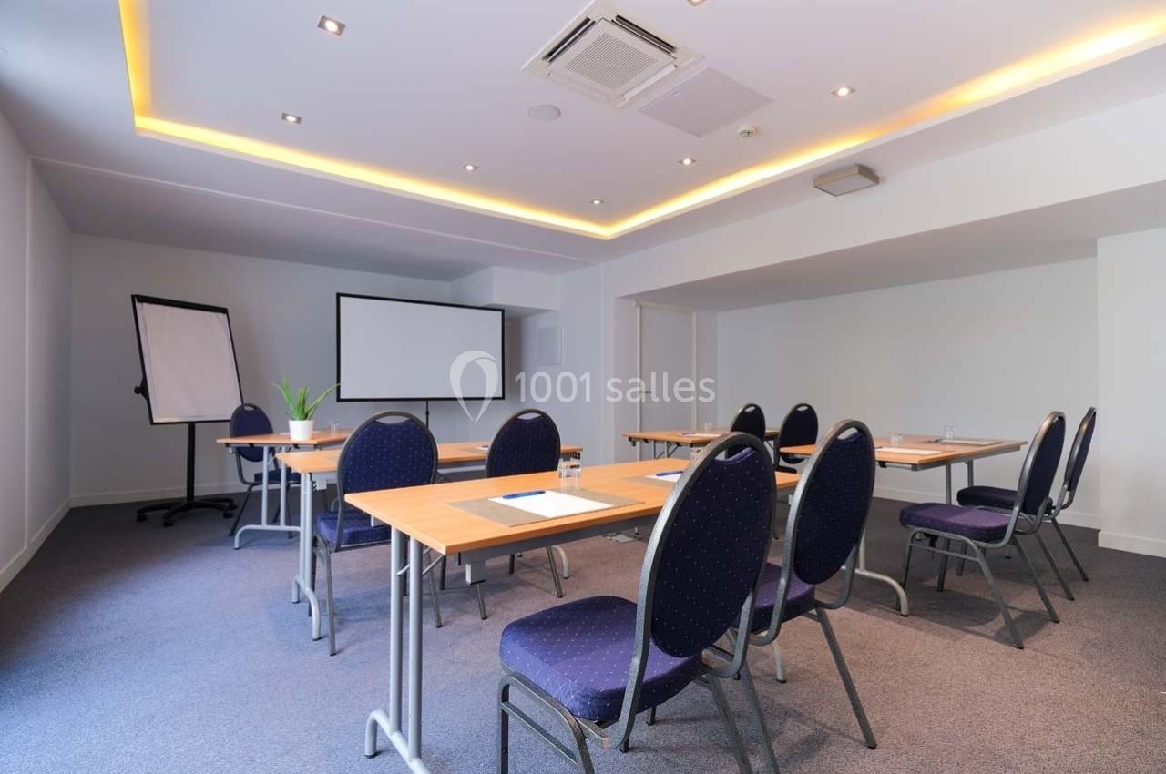 Salle de réunion lumineuse avec tables, chaises, tableau blanc et écran de projection.