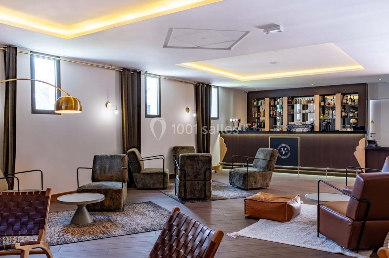 Salon moderne avec fauteuils, tables basses, tapis et un bar éclairé en arrière-plan.