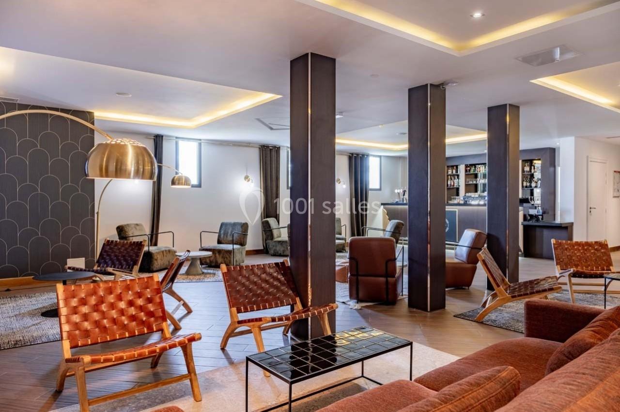 Salon moderne avec fauteuils en cuir tressé, tables basses et éclairage chaleureux, près d'un bar en arrière-plan.