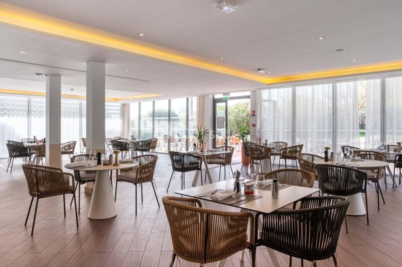 Salle de restaurant lumineuse avec des tables dressées, des chaises en rotin et de grandes baies vitrées.