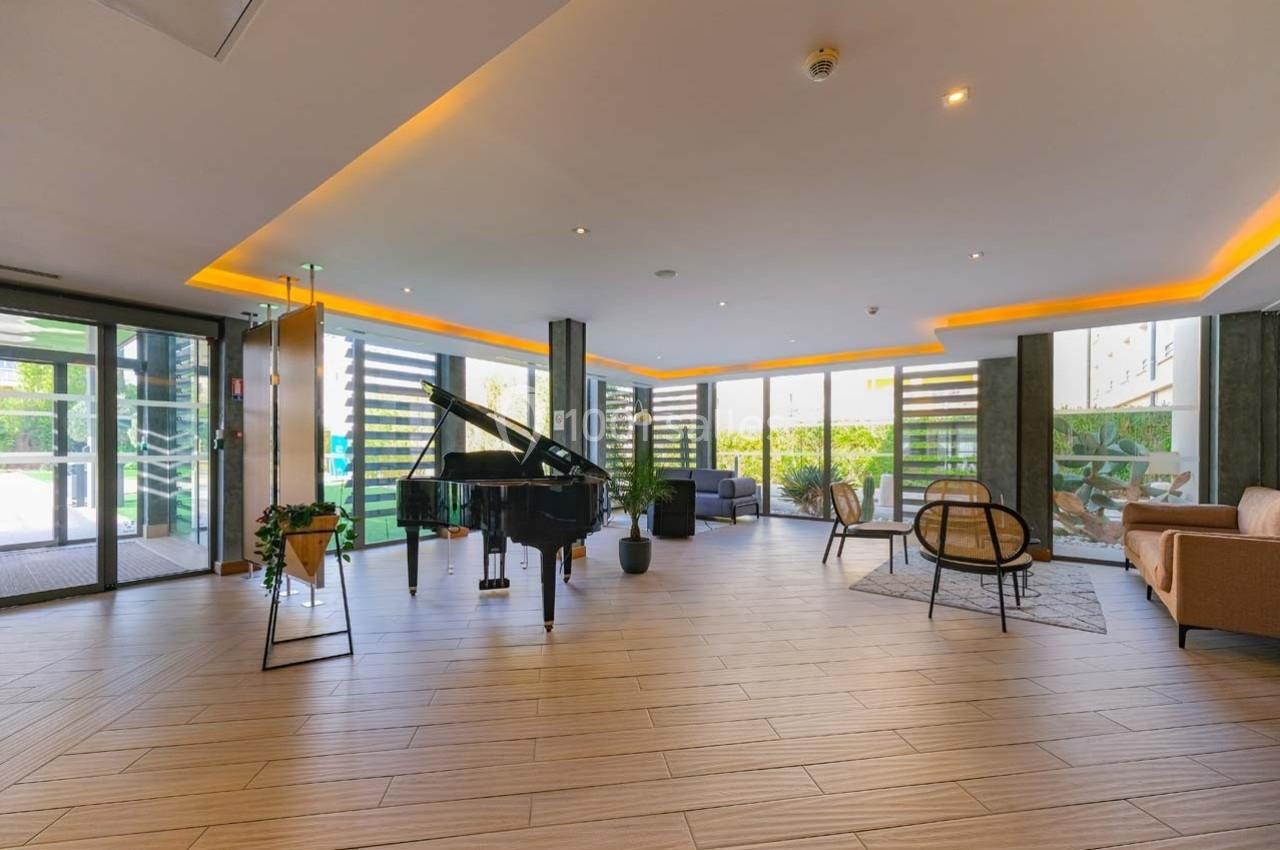 Salon moderne avec un piano à queue, des fauteuils, des plantes et de grandes baies vitrées laissant entrer la lumière…