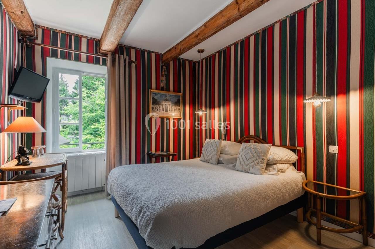 Chambre avec lit double, murs à rayures colorées, poutres apparentes, fenêtre donnant sur un espace vert.