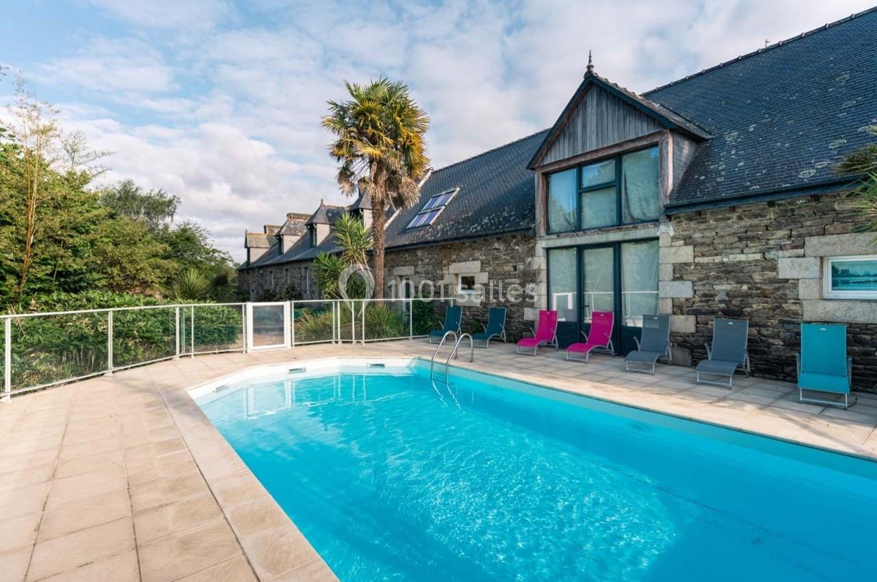 Piscine extérieure entourée de chaises longues devant une maison en pierre avec toit en ardoise.