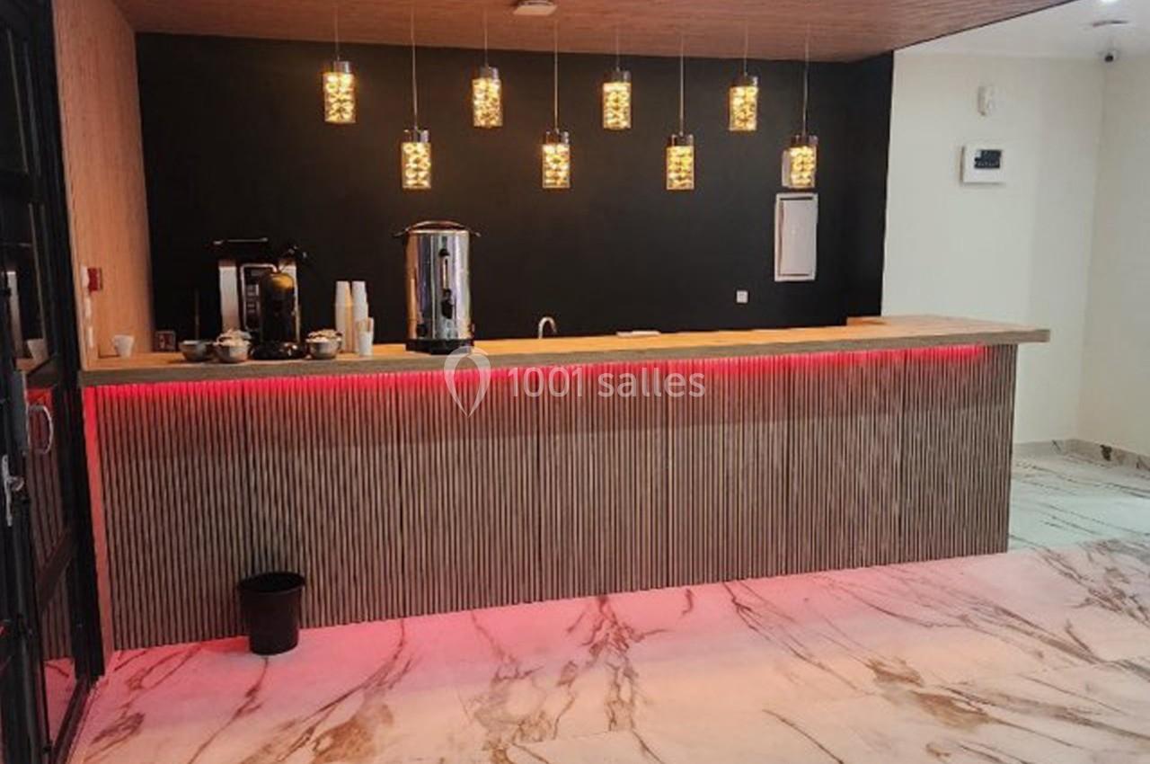 Comptoir en bois éclairé par des suspensions, avec machines à café et sol en marbre dans un espace moderne.