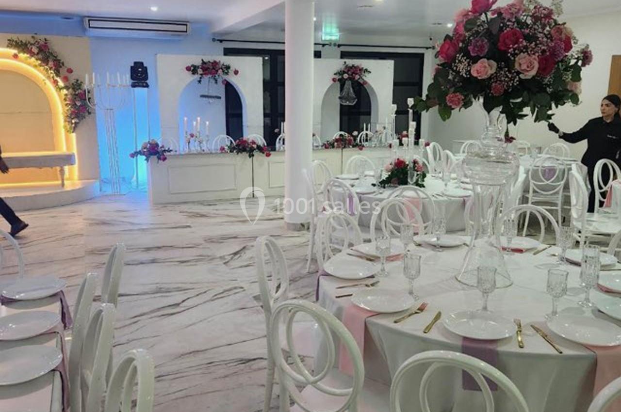 Salle de réception élégante avec tables rondes dressées, chaises blanches et décorations florales roses.