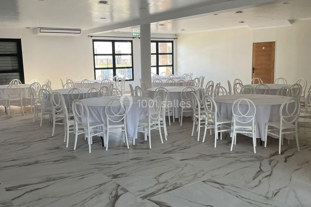 Salle lumineuse avec tables rondes dressées de nappes blanches et chaises blanches sur un sol en marbre clair.