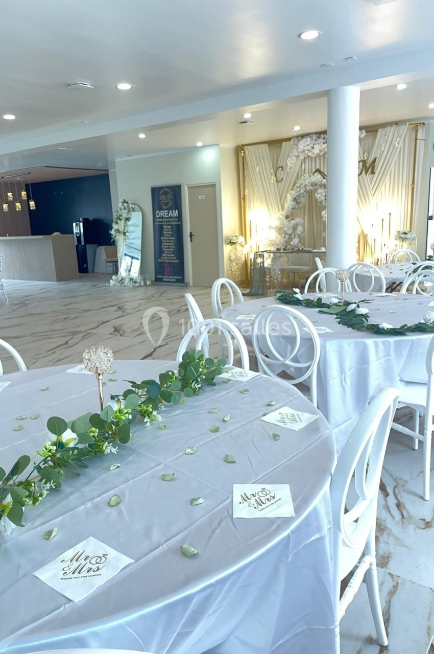 Salle de réception lumineuse avec tables décorées de nappes blanches, feuillages et éléments floraux.
