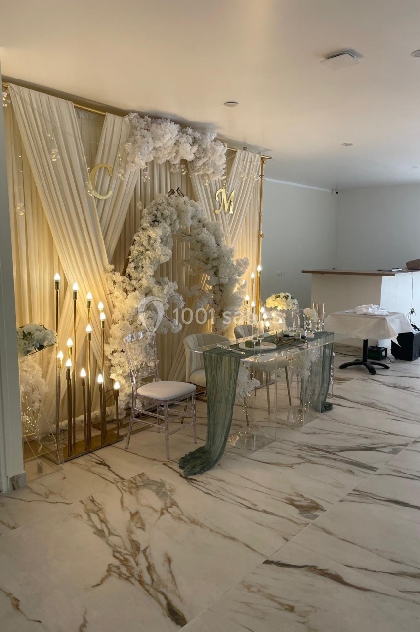 Décoration de mariage élégante avec arche florale blanche, rideaux drapés, guirlandes lumineuses et table transparente.