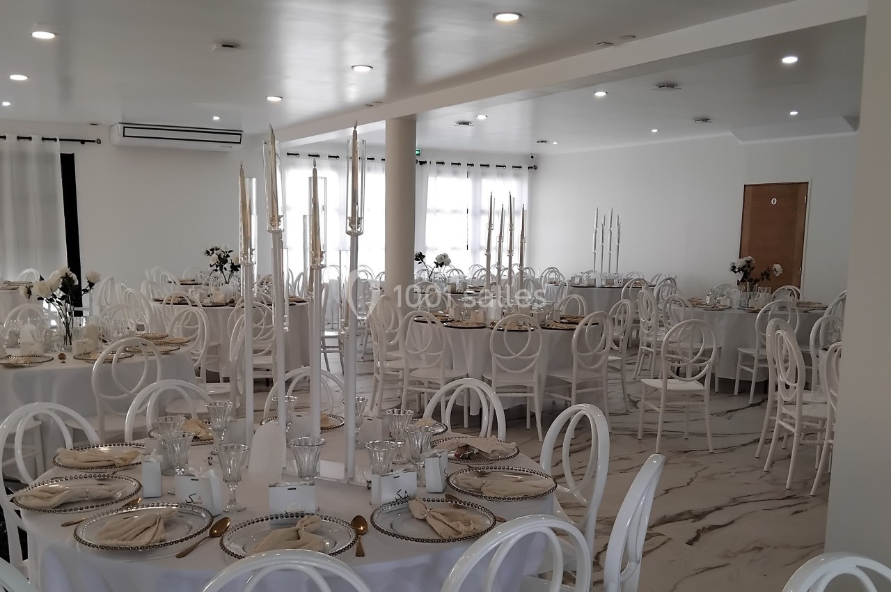Salle de réception élégante avec tables rondes dressées, chaises blanches et chandeliers décoratifs.