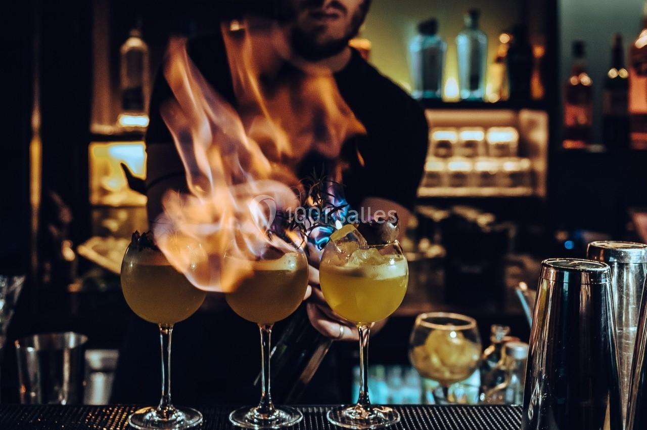Un barman prépare trois cocktails enflammés sur un comptoir dans un bar à l'éclairage tamisé.