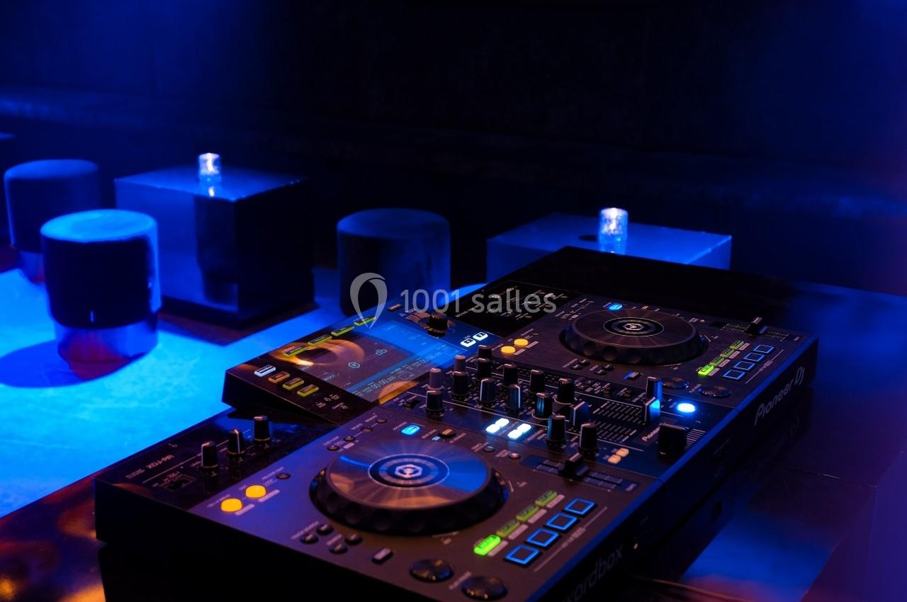 Table de mixage DJ éclairée dans une ambiance sombre, avec des tabourets et tables en arrière-plan.