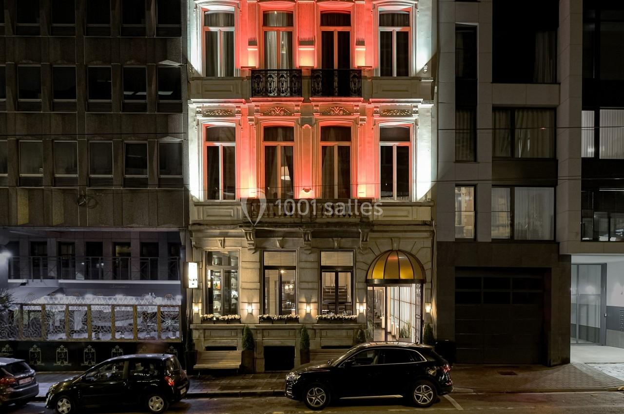 Façade d'un immeuble ancien éclairé en rouge et blanc, entouré de bâtiments modernes, avec des voitures stationnées devant.
