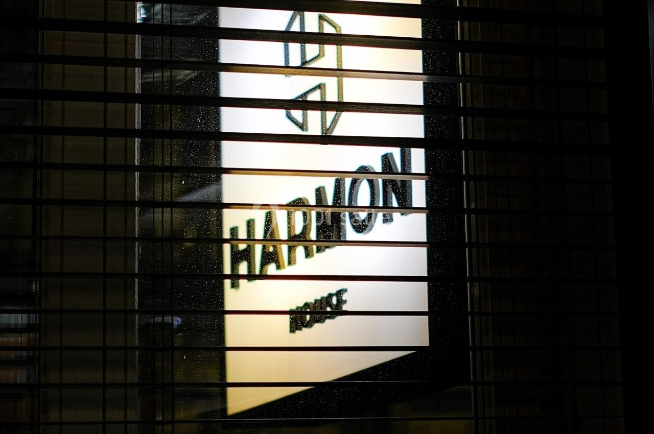 Panneau lumineux avec le logo et le nom ’Harmon’ visible à travers des stores métalliques partiellement ouverts.
