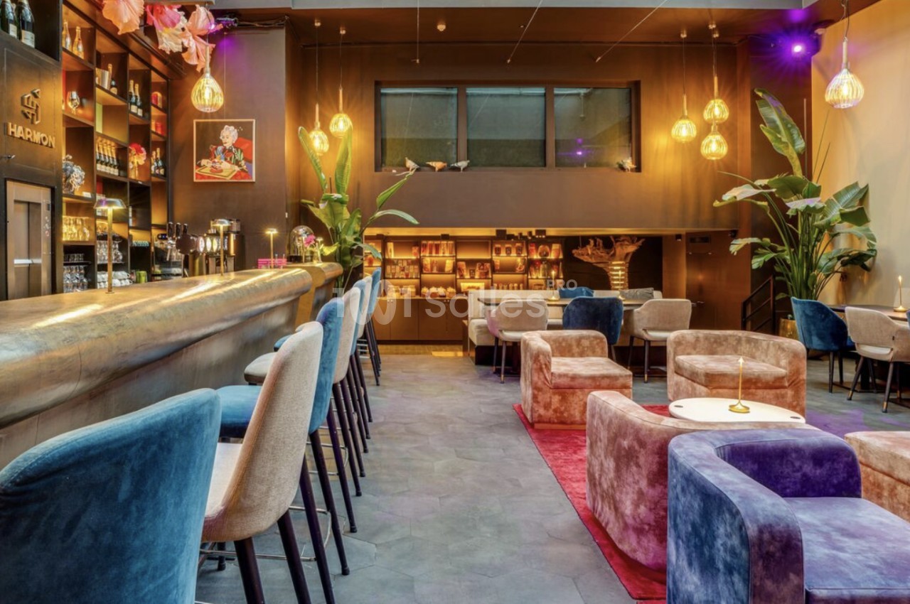 Bar intérieur aux teintes chaudes avec comptoir en métal, tabourets bleus et roses, fauteuils en velours et plantes vertes, éclairé par des suspensions dorées.