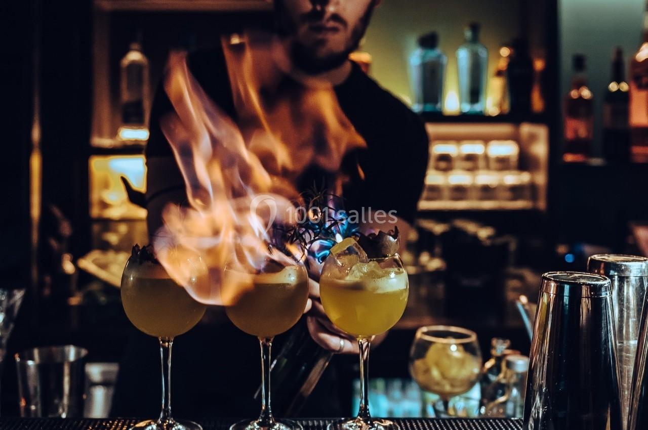 Un barman en train de flamber trois cocktails alignés sur un comptoir dans un bar à l'éclairage tamisé.