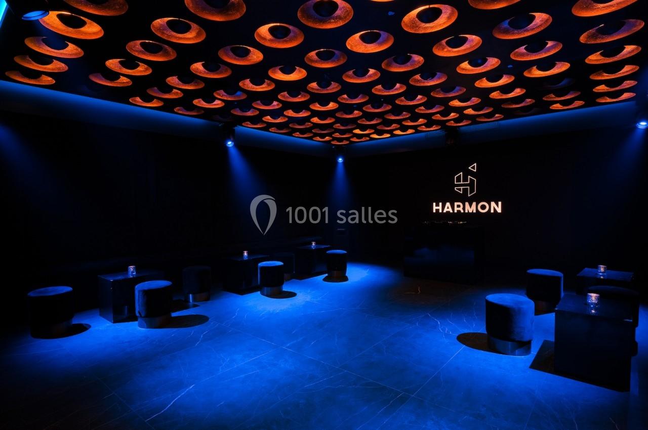Salle sombre avec éclairage bleu, plafond orné de cercles lumineux orange et logo ’HARMON’ illuminé sur un mur.