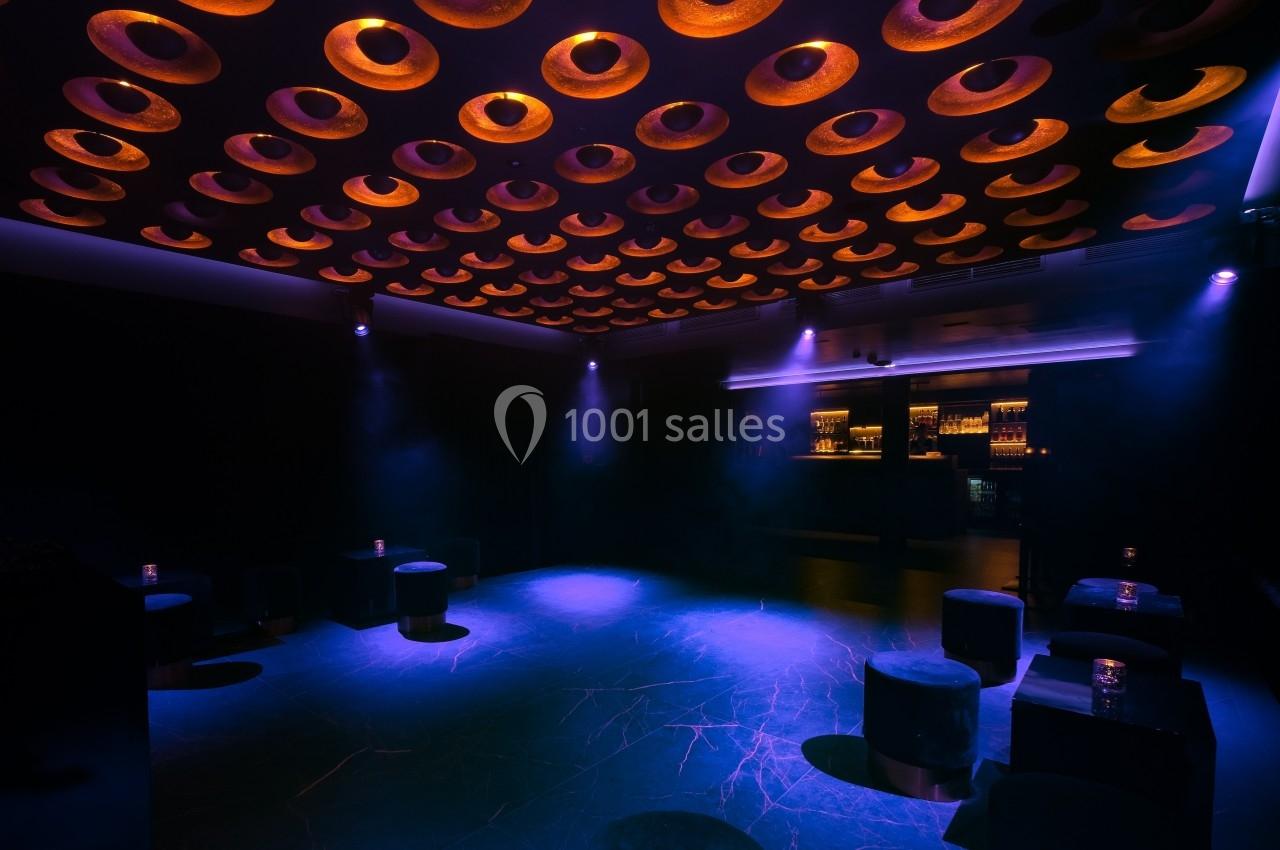Salle sombre avec plafond orné de cercles lumineux, bar éclairé en arrière-plan et sièges bas dispersés.