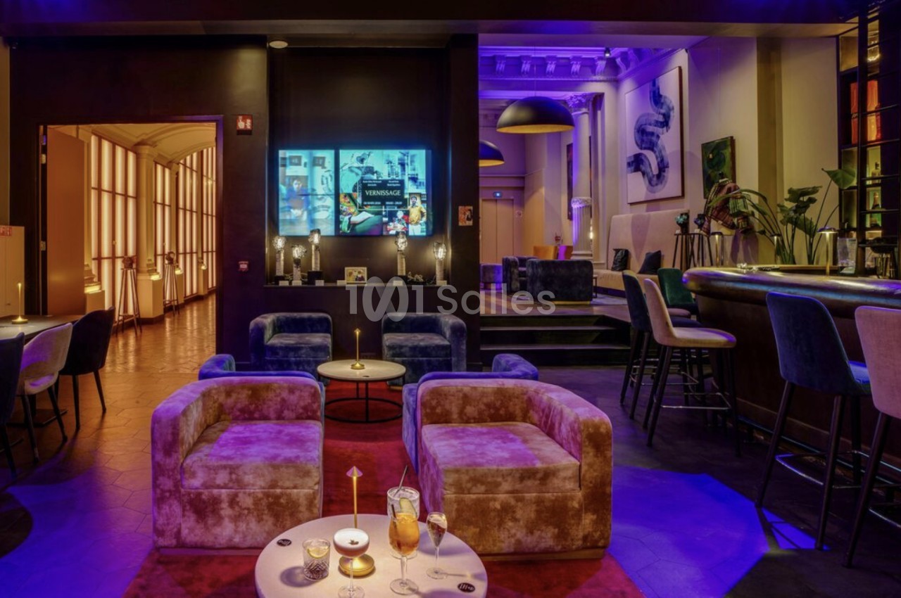 Salon-bar à l'éclairage violet avec fauteuils en velours, tables basses, comptoir en courbe et tabourets, vu depuis un espace voûté aux hautes fenêtres.