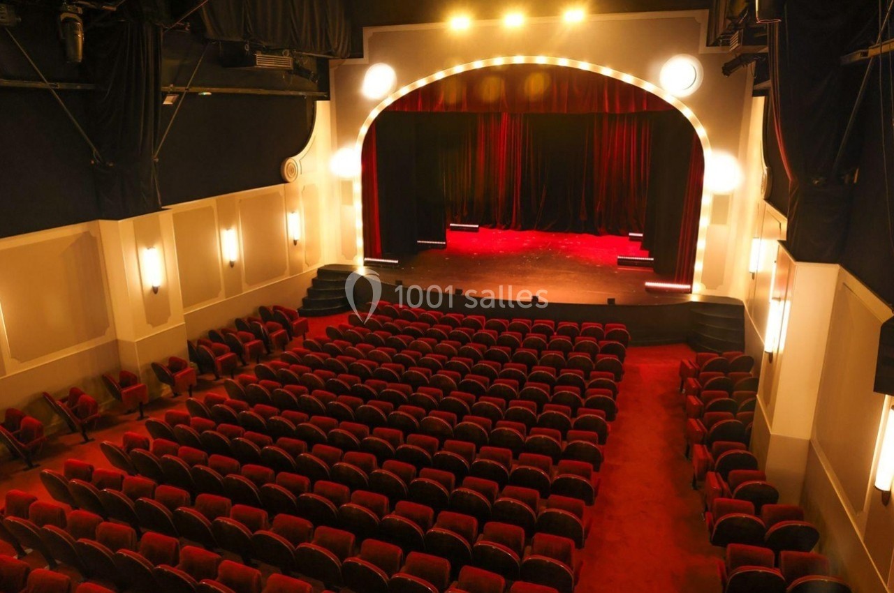 Grande salle du Palais des Glace Salle de théâtre vide avec des rangées de sièges rouges, une scène éclairée et un rideau rouge fermé.