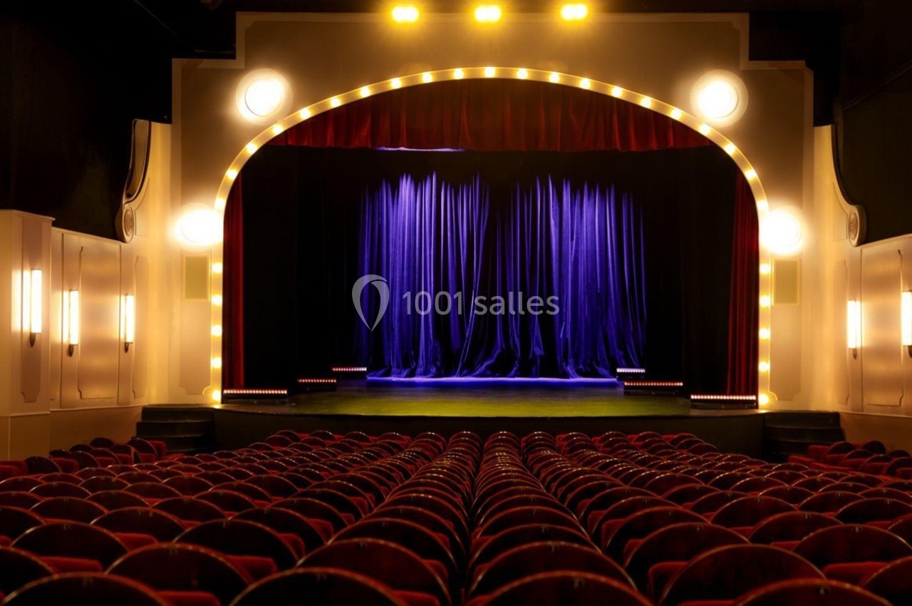 Grande salle du Palais des Glaces Salle de théâtre vide avec des rangées de sièges rouges et une scène éclairée ornée de rideaux bleus.