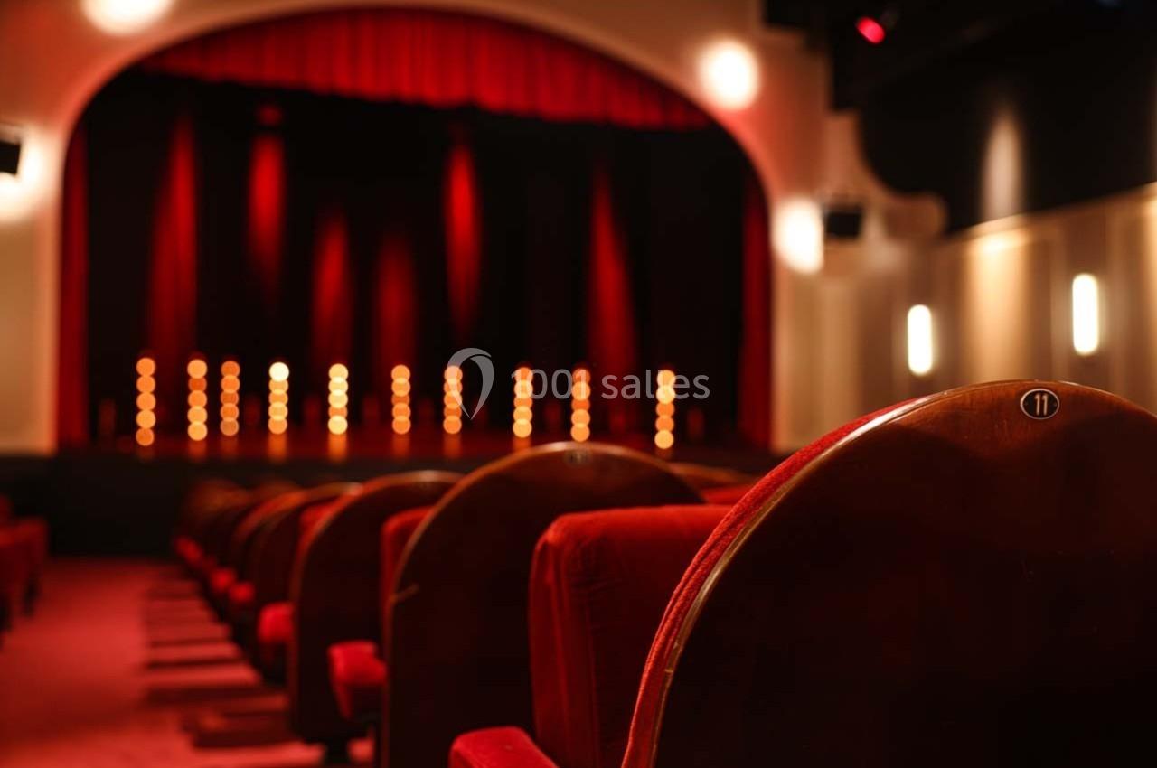 Grande salle du Palais des Glaces Salle de théâtre avec des sièges rouges au premier plan et une scène éclairée par des lumières en arrière-plan.