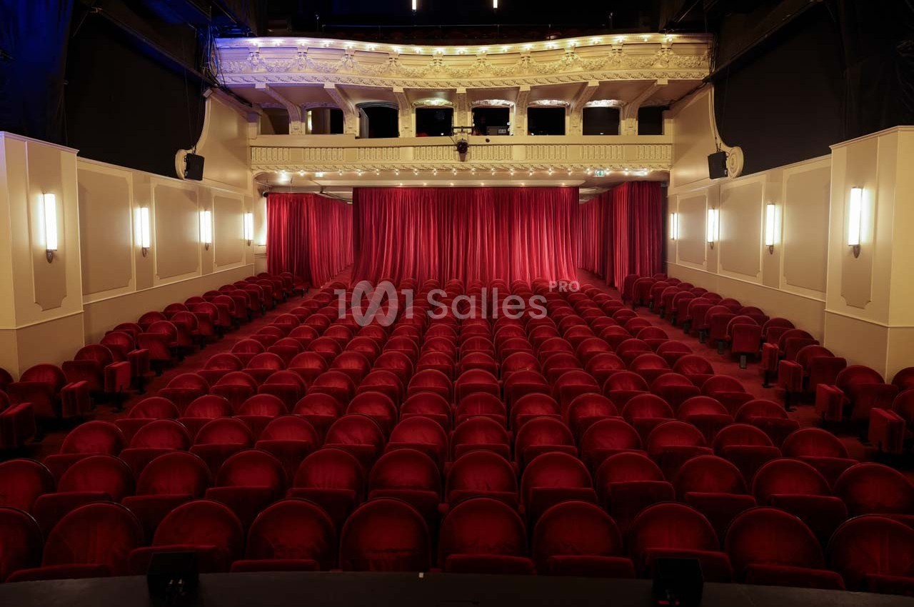 Salle de théâtre vide avec des rangées de fauteuils rouges et une scène fermée par un rideau rouge.