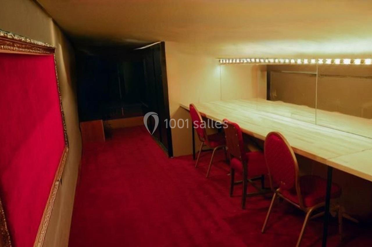 Loge arrière scène Salle avec moquette rouge, table en bois clair, chaises rouges et miroir éclairé par des ampoules.