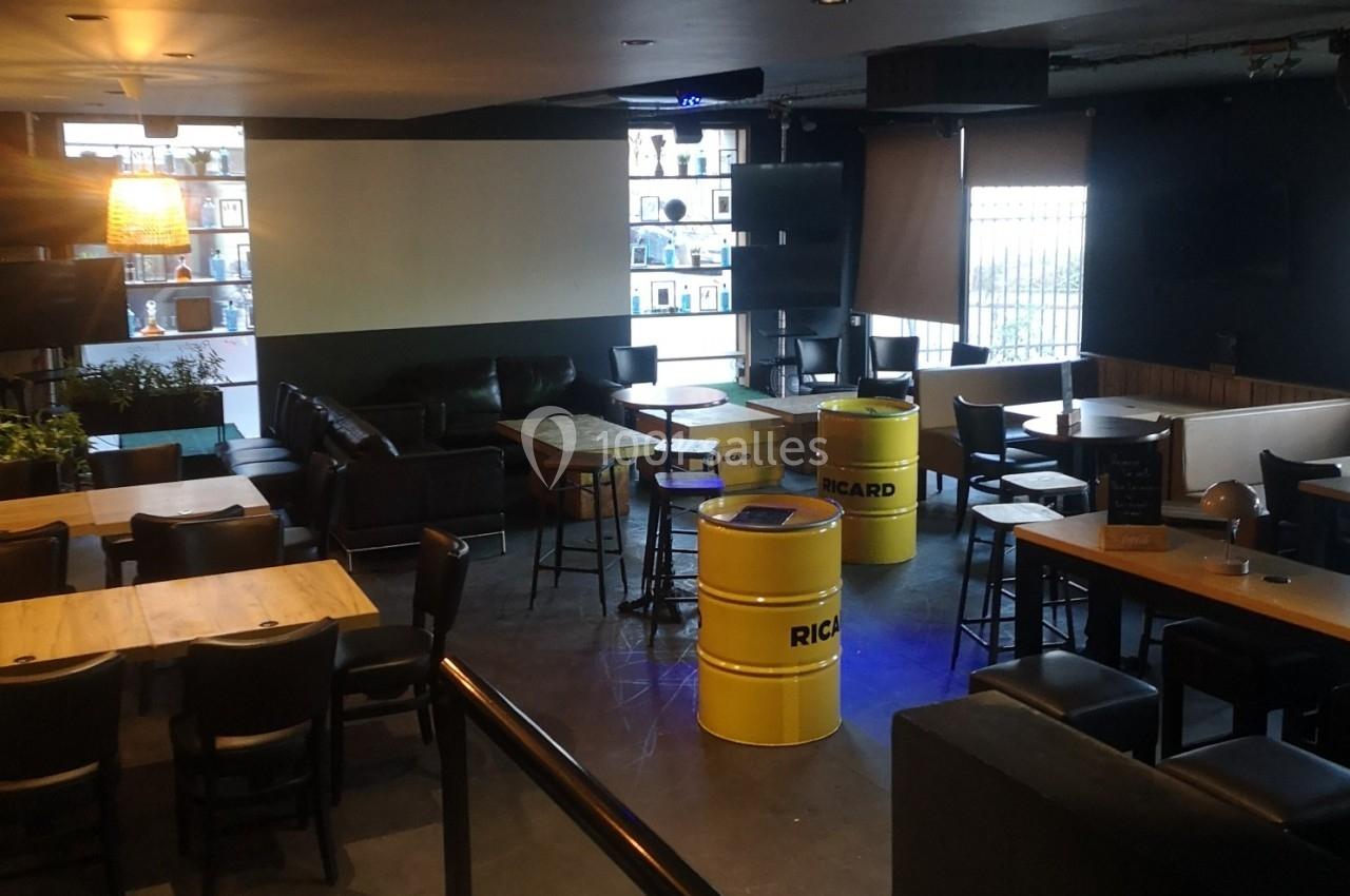 Salle de bar avec tables en bois, chaises noires, tonneaux jaunes en guise de tables et éclairage tamisé.