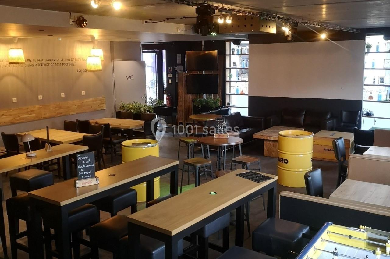 Salle de bar moderne avec tables en bois, fûts recyclés comme mobilier, canapé, baby-foot et éclairage tamisé.