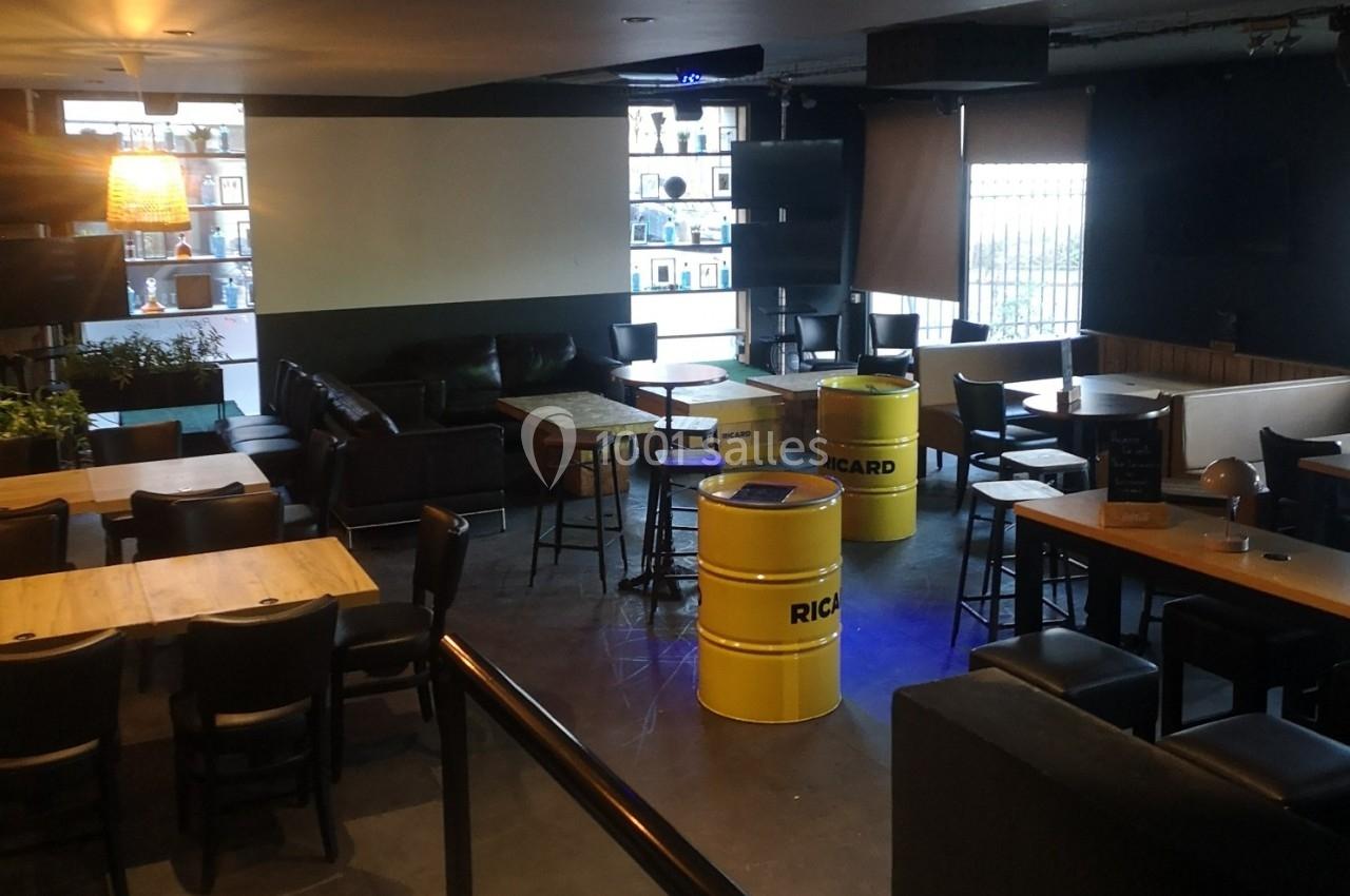 Salle de bar avec tables en bois, chaises noires, tonneaux jaunes Ricard et canapés dans une ambiance tamisée.