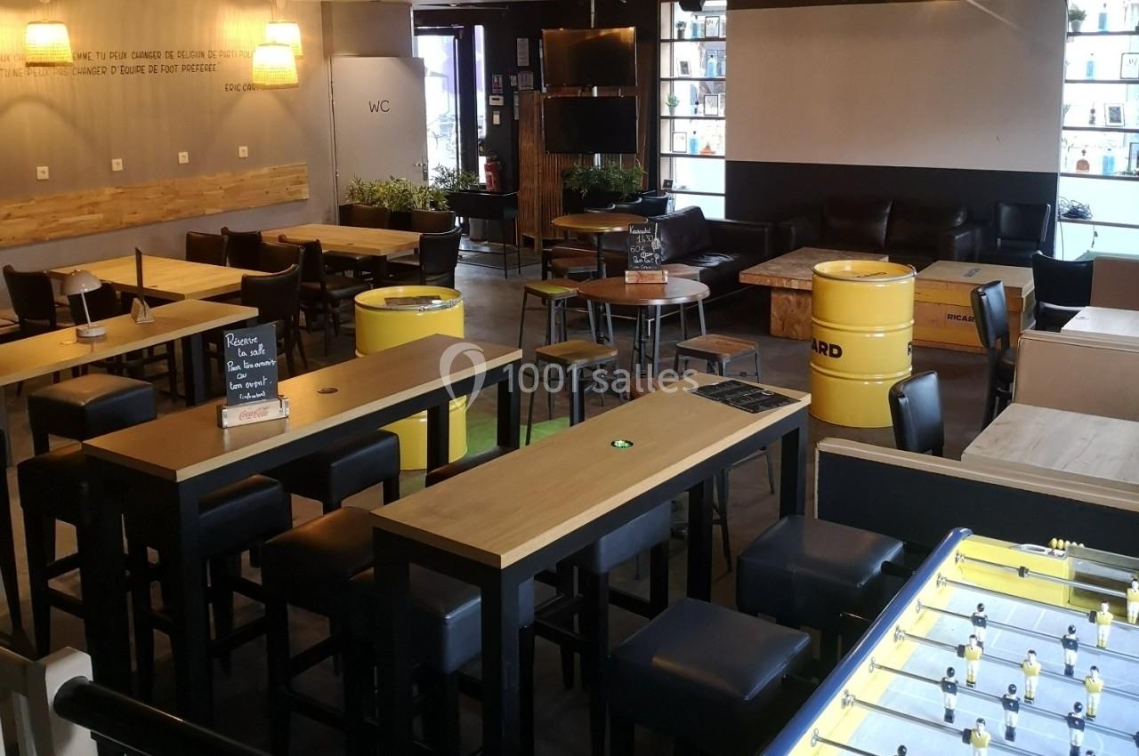 Salle de bar moderne avec tables hautes, tabourets, baby-foot et tonneaux jaunes servant de décoration.